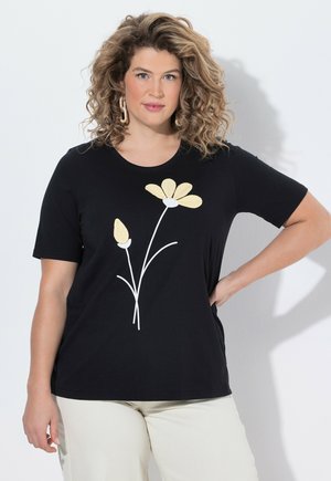 Kobieta z kręconymi włosami, ubrana w czarny T-shirt z żółtym wzorem kwiatu i jasne spodnie, pozująca z jedną ręką na biodrze.