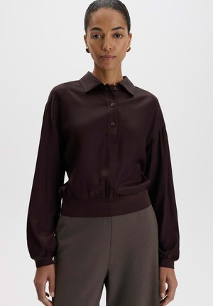 Vrouw draagt een donkerbruine blouse met lange mouwen en kraag en grijze hoge taille broek tegen een effen witte achtergrond.