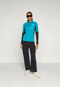 Γυναίκα που φοράει μπλε ζακέτα με φερμουάρ, μαύρο cargo παντελόνι, λευκά αθλητικά παπούτσια και μεγάλα σκούρα γυαλιά ηλίου, στέκεται μπροστά σε ένα απλό ανοιχτό φόντο.