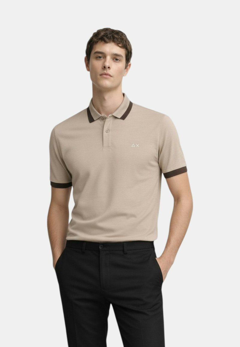 Giovane uomo che indossa una polo beige con rifiniture scure sul colletto e sulle maniche, abbinata a pantaloni neri, mani nelle tasche, espressione neutra.