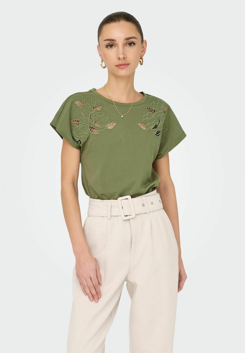 T-shirt vert olive avec broderies florales et détails découpés, associé à un pantalon taille haute beige clair sur mesure et une ceinture.