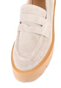 Chaussure beige en daim à enfiler avec une bride rembourrée, des coutures discrètes et une semelle épaisse en gomme. Texture douce et forme arrondie.