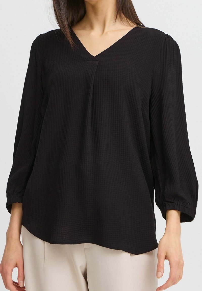 Blouse noire avec un col en V, tissu texturé et manches 3/4. Présente des poignets froncés et une coupe décontractée. Associée à un pantalon de couleur claire.