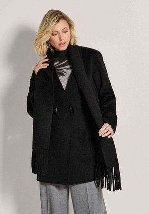 Vrouw met blond haar die een zwarte jas, zwarte sjaal met franjes, grijze gebreide trui met patroon en lichtgrijze broek draagt tegen een effen achtergrond.