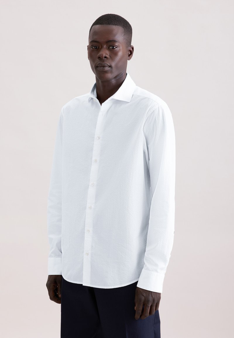 Homme aux cheveux courts portant une chemise blanche à manches longues boutonnée et un pantalon foncé, debout devant un fond clair uni.