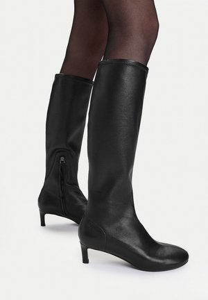 Knielange schwarze Lederstiefel mit niedrigen Absätzen, getragen über schwarzen transparenten Strumpfhosen, aus Seiten- und Rückansichten vor weißem Hintergrund gezeigt.