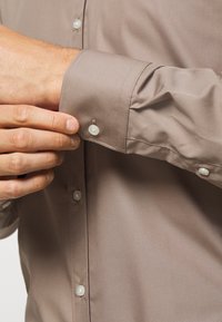 Brown dress shirt featuring a smooth fabric, button-up front with visible pearly buttons, and a fitted cuff design.