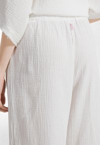 Lähikuva henkilöstä, joka käyttää valkoisia teksturoituja housuja, joissa on joustava vyötärönauha ja pieni vaaleanpunainen DEHA-labeli takana.