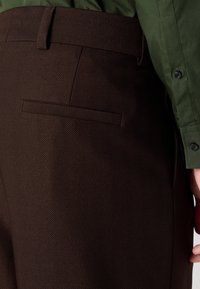 Pantalons en chevron marron avec un devant plat et une poche arrière. Associés à une chemise verte, mettant en avant une texture douce et une coupe ajustée.