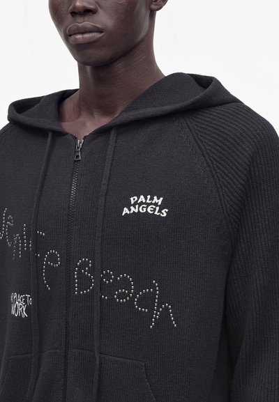 Μαύρο φούτερ με φερμουάρ, κεντημένο με το "Palm Angels" στο στήθος και διακοσμημένο με κείμενο από στρας μπροστά, φορεμένο από άτομο με σκούρο δέρμα.