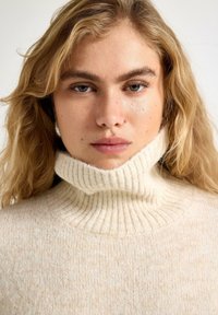 Kremfarvet rullekravesweater lavet af blødt, tekstureret stof med ribdetaljer ved kraven og ærmekanterne. Enkel, hyggelig design.