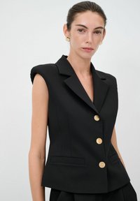 Schwarzer ärmelloser Blazer aus strukturiertem Stoff mit Reverskragen, goldenen Knöpfen und seitlichen Taschen, taillierte Passform, glatte Textur.