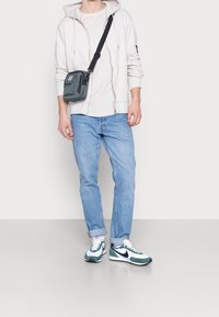 Světle šedá mikina na zip s předními kapsami, bílý tričko, modré džíny a zeleno-bílé tenisky Nike. Šedá kabelka přes rameno se zipem.