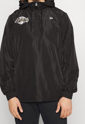 Mann trägt schwarze Kapuzenjacke mit Halb-Reißverschluss, elastischen Bündchen, Los Angeles Lakers-Logo auf der linken Brust und einem kleinen weißen Logo auf der rechten Brust.