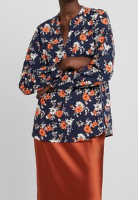 Blusa floreale a maniche lunghe in blu navy con fiori arancioni e bianchi, dotata di colletto alla coreana e una vestibilità dritta con spacchi laterali.