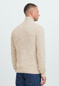 Mann trägt einen beigen Rollkragenpullover und blaue Jeans, steht seitlich mit dem Rücken größtenteils zur Kamera vor einem weißen Hintergrund.