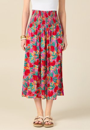Kobieta ubrana w kolorową, midi spódnicę o wysokim stanem z kwiatowym wzorem w odcieniach czerwieni, zieleni, różu i pomarańczy, w połączeniu z białym topem i sandałami.