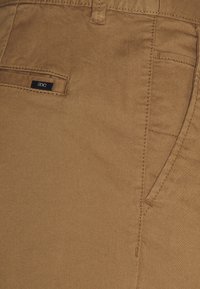 Braune Baumwollhose mit glatter Textur und Seitentaschen. Verfügt über ein kleines schwarzes Etikett nahe der Gesäßtasche. Einfaches Design mit klaren Linien.