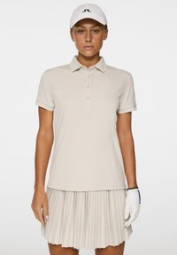 Beżowa koszulka polo z krótkim rękawem, z zapięciem na guziki, oraz plisowana spódnica. Biała rękawica golfowa i czapka z logo.