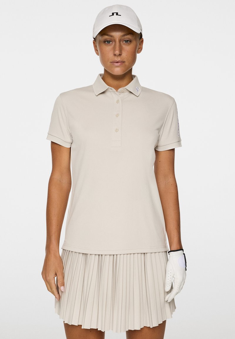 Beżowa koszulka polo z krótkim rękawem, z zapięciem na guziki, oraz plisowana spódnica. Biała rękawica golfowa i czapka z logo.