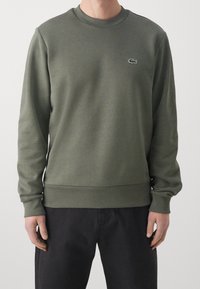 Uomo che indossa una felpa a maniche lunghe color verde oliva con colletto girocollo e un piccolo logo di coccodrillo sul lato sinistro del petto, abbinata a pantaloni neri.