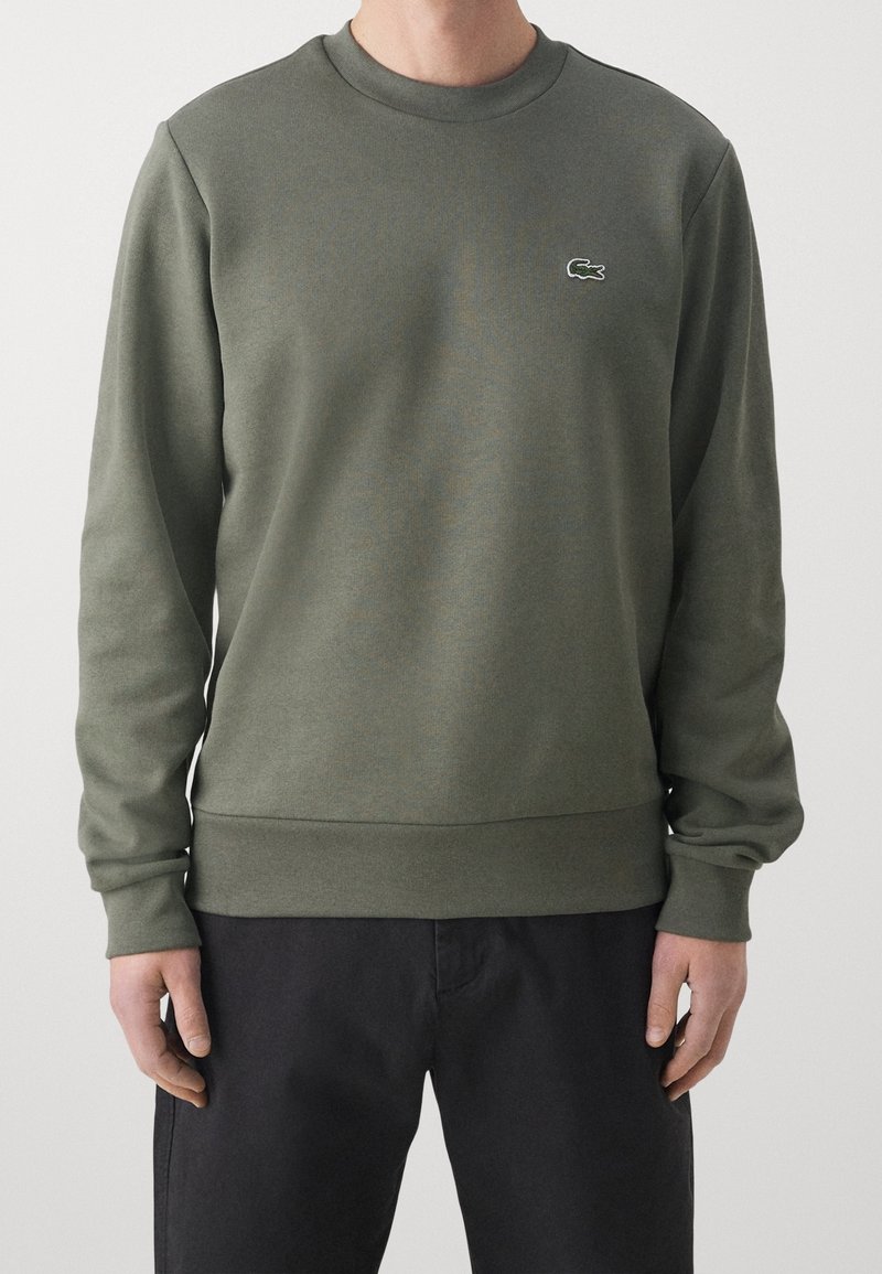 Uomo che indossa una felpa a maniche lunghe color verde oliva con colletto girocollo e un piccolo logo di coccodrillo sul lato sinistro del petto, abbinata a pantaloni neri.