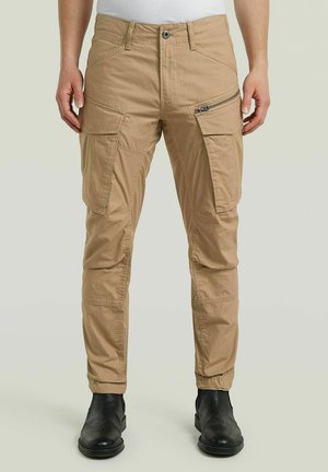 Pantalon cargo beige avec plusieurs poches, dont une poche zippée sur la cuisse, porté avec des bottines noires sur un fond neutre.