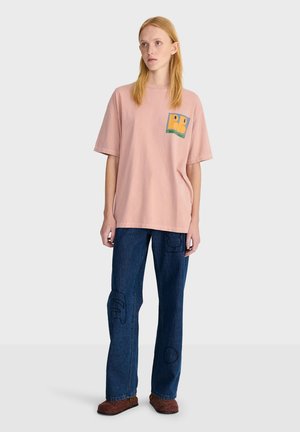 Jeune femme debout portant un t-shirt rose ample avec un petit motif, un jean bleu foncé avec des patchs cousus et des chaussures marron.