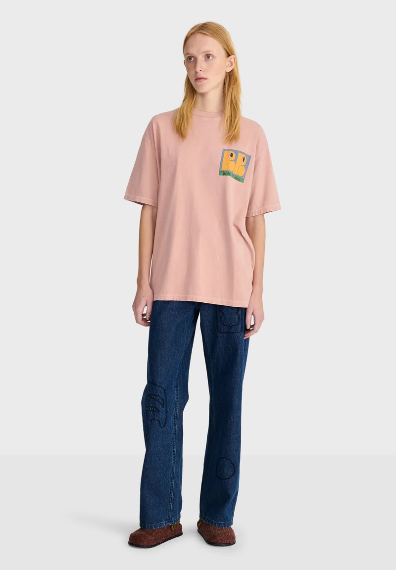 Jeune femme debout portant un t-shirt rose ample avec un petit motif, un jean bleu foncé avec des patchs cousus et des chaussures marron.