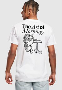 Weißes Baumwoll-T-Shirt mit einer schwarzen Cartoon-Illustration einer Person, die eine Zeitung liest, mit dem Text "Die Kunst der Morgen" darüber.