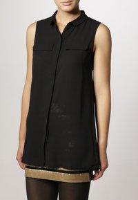 Utan ärmar svart knäppt blus med två bröstfickor, bärs över en guldsequinskjol och svarta strumpbyxor.