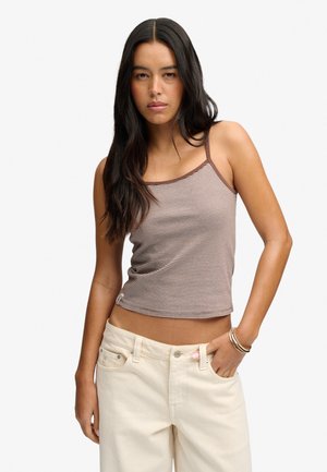 Kvinde med langt mørkt hår iført en ærmeløs brun og hvid stribet top og lyse beige bukser, med den ene hånd i lommen.