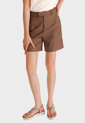 Donna che indossa pantaloncini marroni al ginocchio, top bianco e sandali beige piatti, in piedi davanti a uno sfondo chiaro uniforme.