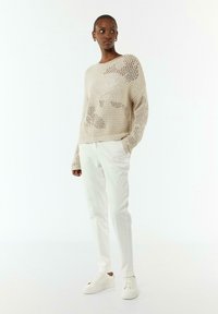 Beiger Strickpullover mit lockerer Passform, der ein großes Netzmuster aufweist. Kombiniert mit maßgeschneiderten weißen Hosen und weißen Sneakern.