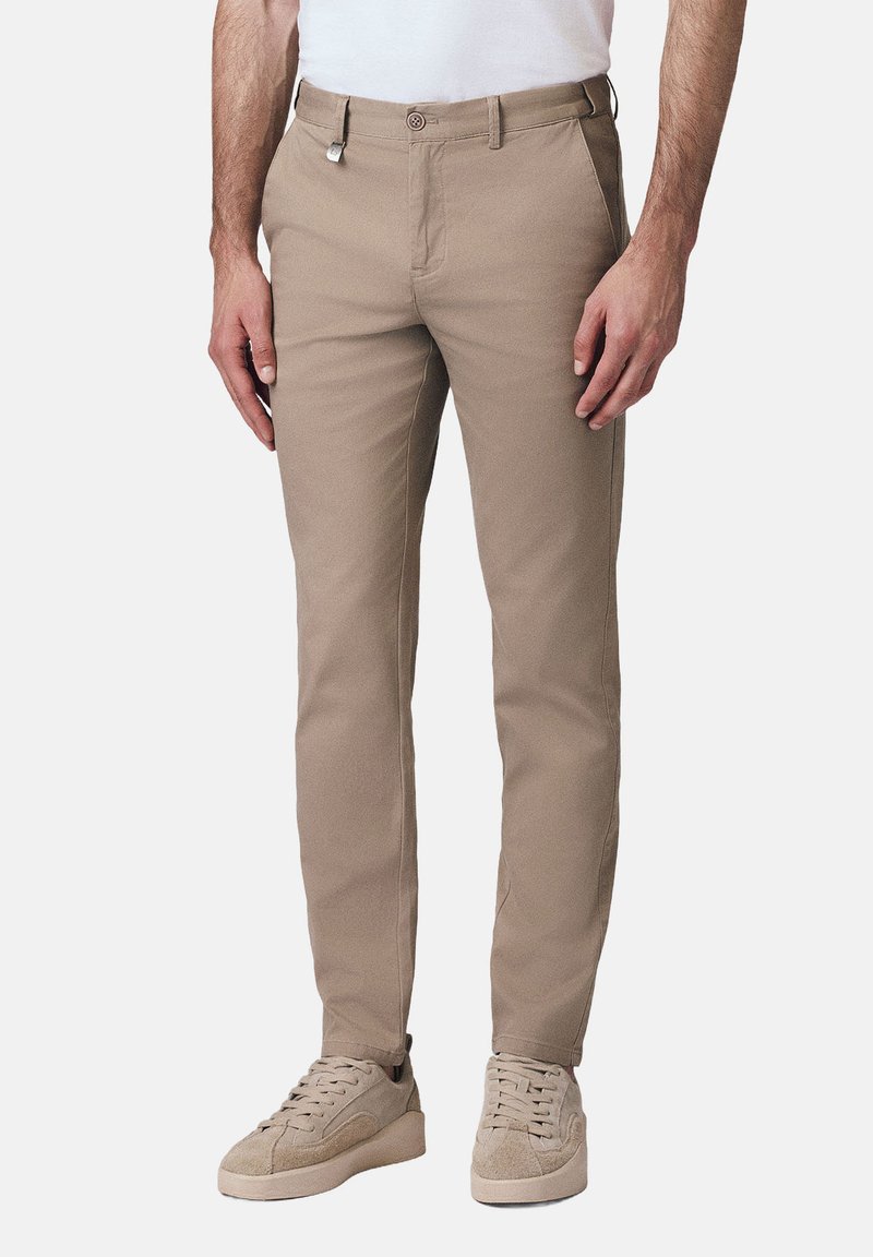 Mężczyzna w beżowych dopasowanych spodniach i pasujących beżowych trampkach, stojący na tle gładkiej białej ściany.