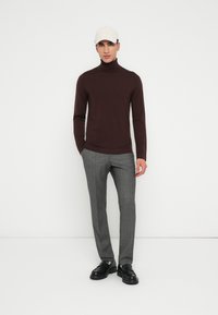 Bărbat îmbrăcat cu o șapcă bej, un pulover negru cu guler înalt, pantaloni gri eleganți și pantofi de piele negri, stând în fața unui fundal alb.