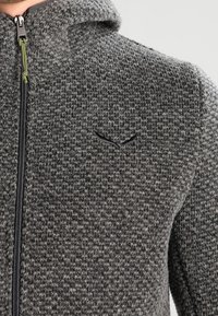 Szary dzianinowy kurtka z kapturem z teksturowanym wzorem, czarnym zamkiem błyskawicznym i subtelnym logo na piersi. Miękki materiał o swobodnym kroju.