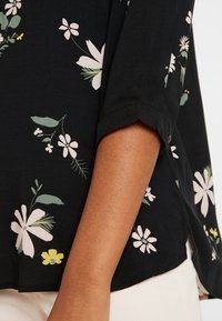 Blouse noire à manches retroussées, avec un motif floral en blanc, vert et jaune. Tissu doux et détail d'ourlet arrondi visible.