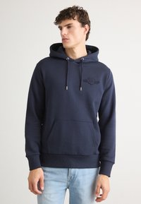 Námornícky modrá mikina s kapucňou vyrobená z mäkkej tkaniny, s prednou kapsou, šnúrkou na kapucni a embosovaným logom na hrudi.