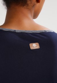 Navyblaues T-Shirt mit weitem Ausschnitt, ausgestattet mit einem gestreiften Saum und einem hellbraunen Lederlabel mit geprägt Logo auf der Rückseite. Glatte Textur.