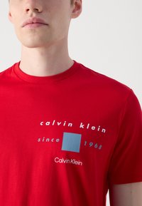 Punane puuvillane T-särk, millel on ümmargune kaelus, valge ja sinise graafikatekstiga, sealhulgas "calvin klein since 1968" ja sinine ruut.