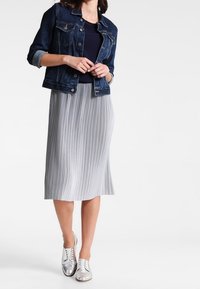 Farkkutakki rintataskuilla, yhdistettynä harmaaseen laahustavaan midi-hameeseen ja hopeisiin lenkkareihin, joissa on pyöreä kaula-aukko ja lyhyet hihat.