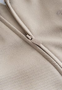 Beige Kleidungsstück mit Reißverschluss und strukturierter Stoffoberfläche. Nahaufnahme des Reißverschluss-Zugs mit der Aufschrift "BAZAR", die das Design und die Materialdetails hervorhebt.