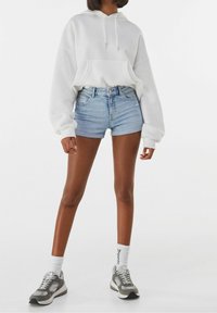 Vit hoodie med framficka, matchad med ljusblå denimshorts. Modellen bär grå sneakers med vita snören och knästrumpor.