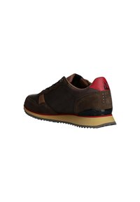 Sneaker in suede marrone con accenti rossi, tallone nero e suola beige. Presenta un sistema di allacciatura e una parte posteriore testurizzata con dettagli a ventilazione circolari.