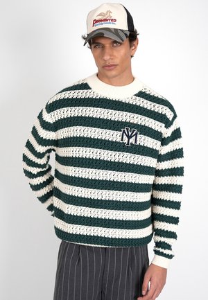 Homme portant un pull en maille rayé vert et blanc avec un emblème, un pantalon à fines rayures et une casquette de camionneur beige avec un logo.