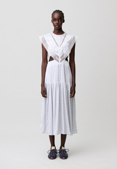 Donna che indossa un vestito bianco con fantasia, dettagli in pizzo, lati ritagliati e sneakers blu scuro, in piedi davanti a uno sfondo grigio chiaro uniforme.