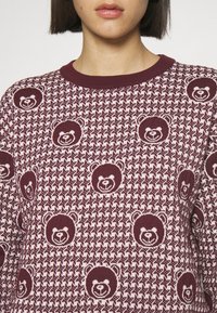 Femme portant un pull bordeaux et blanc avec un motif pied-de-poule et des motifs répétitifs de visages d’ours souriants.