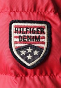 Rød stof med et broderet skjoldformet mærke. Mærket har vandrette striber, "HILFIGER DENIM", og fem stjerner i marineblå og hvid.