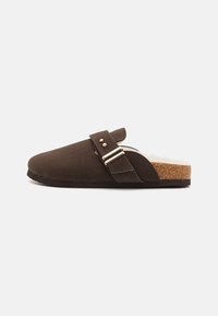 Brune semskede klosser har en spenne, myk innersåle og en korksåle. Designet er minimalistisk med en jevn overflate og rund tå.